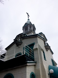 A historic church with light-colored stone walls and green accents. It features a tall, pointed steeple topped with a cross. Statues are present in recessed areas of the structure, highlighting its religious significance. Architectural details include arched windows and decorative cornices.