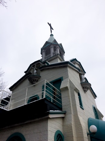 A historic church with light-colored stone walls and green accents. It features a tall, pointed steeple topped with a cross. Statues are present in recessed areas of the structure, highlighting its religious significance. Architectural details include arched windows and decorative cornices.