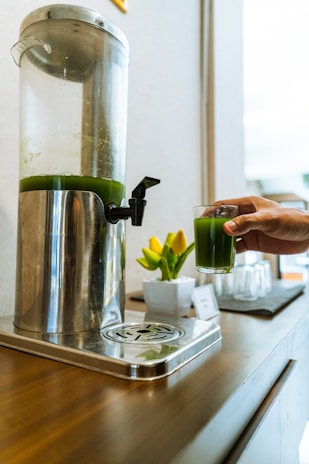 A hand holding a glass of green juice being poured from a large, metallic dispenser situated on a wooden surface. A small pot with bright yellow tulips sits nearby, adding a touch of color. Glasses and a note card are also visible in the background, creating a refreshing and inviting scene.