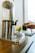 A hand holding a glass of green juice being poured from a large, metallic dispenser situated on a wooden surface. A small pot with bright yellow tulips sits nearby, adding a touch of color. Glasses and a note card are also visible in the background, creating a refreshing and inviting scene.