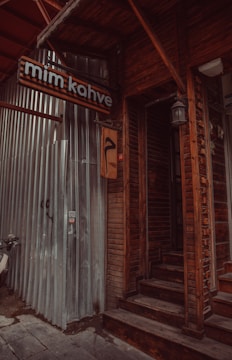 A rustic wooden entrance to a building with a sign that reads 'mim kahve'. The exterior combines wooden panels and corrugated metal. A motorcycle is partially visible on the left side. Dim lighting and shadows create an intimate atmosphere.