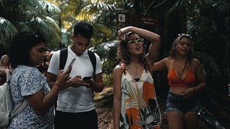 A group of young people are walking through a lush, tropical forest. One person is using a smartphone, while another seems to be adjusting their glasses. Dense green foliage surrounds them, creating a vibrant natural backdrop.