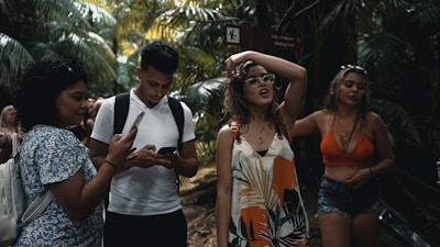 A group of young people are walking through a lush, tropical forest. One person is using a smartphone, while another seems to be adjusting their glasses. Dense green foliage surrounds them, creating a vibrant natural backdrop.