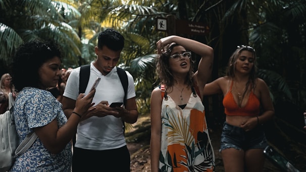 A group of young people are walking through a lush, tropical forest. One person is using a smartphone, while another seems to be adjusting their glasses. Dense green foliage surrounds them, creating a vibrant natural backdrop.