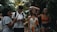 A group of young people are walking through a lush, tropical forest. One person is using a smartphone, while another seems to be adjusting their glasses. Dense green foliage surrounds them, creating a vibrant natural backdrop.