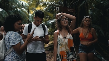 A group of young people are walking through a lush, tropical forest. One person is using a smartphone, while another seems to be adjusting their glasses. Dense green foliage surrounds them, creating a vibrant natural backdrop.