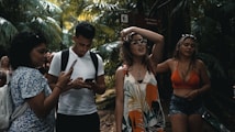 A group of young people are walking through a lush, tropical forest. One person is using a smartphone, while another seems to be adjusting their glasses. Dense green foliage surrounds them, creating a vibrant natural backdrop.