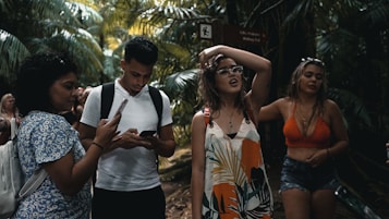 A group of young people are walking through a lush, tropical forest. One person is using a smartphone, while another seems to be adjusting their glasses. Dense green foliage surrounds them, creating a vibrant natural backdrop.