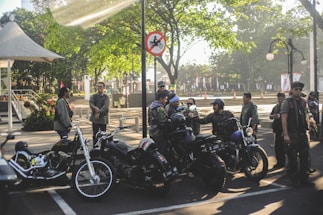 A group of people in casual outdoor attire are gathered around several parked motorcycles in an urban park setting. The area is shaded by trees and a nearby gazebo, with sunlight filtering through leaves. A sign nearby prohibits motorcycles in a certain area. The atmosphere is social and relaxed.