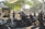 A group of people in casual outdoor attire are gathered around several parked motorcycles in an urban park setting. The area is shaded by trees and a nearby gazebo, with sunlight filtering through leaves. A sign nearby prohibits motorcycles in a certain area. The atmosphere is social and relaxed.