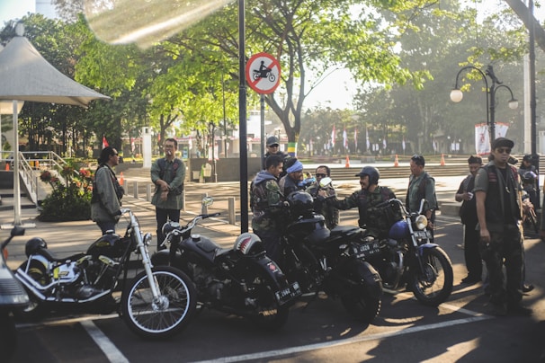A group of people in casual outdoor attire are gathered around several parked motorcycles in an urban park setting. The area is shaded by trees and a nearby gazebo, with sunlight filtering through leaves. A sign nearby prohibits motorcycles in a certain area. The atmosphere is social and relaxed.