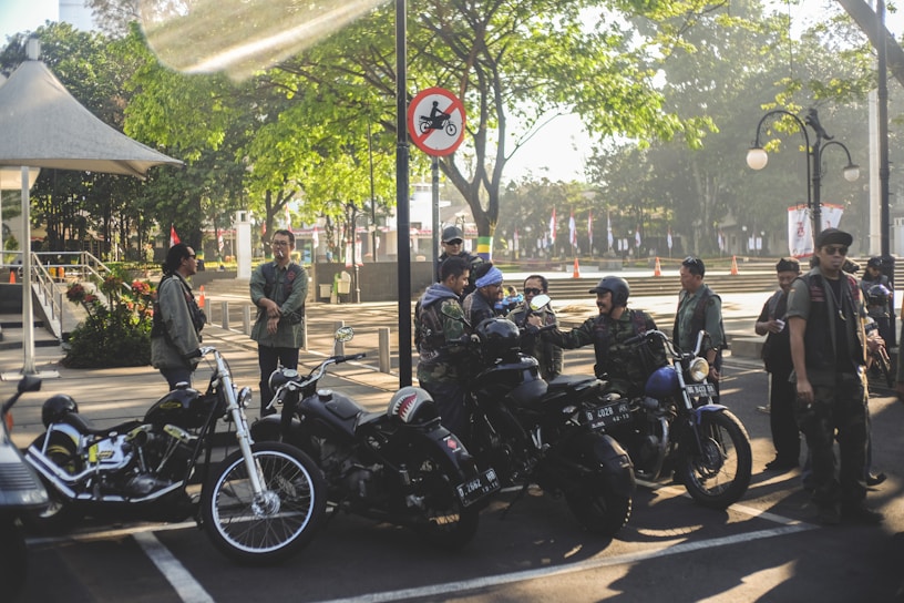 A group of people in casual outdoor attire are gathered around several parked motorcycles in an urban park setting. The area is shaded by trees and a nearby gazebo, with sunlight filtering through leaves. A sign nearby prohibits motorcycles in a certain area. The atmosphere is social and relaxed.
