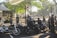 A group of people in casual outdoor attire are gathered around several parked motorcycles in an urban park setting. The area is shaded by trees and a nearby gazebo, with sunlight filtering through leaves. A sign nearby prohibits motorcycles in a certain area. The atmosphere is social and relaxed.