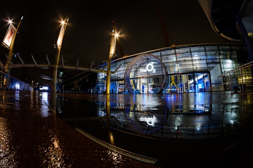 A modern architectural structure with glass panels and a large circular entrance displaying 'The O2' is illuminated by artificial lighting. The scene is reflected on a wet surface, enhancing the visual symmetry. Yellow poles with lights flank the entrance, contributing to a futuristic ambiance.