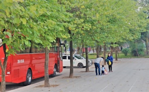 A red bus is parked along a tree-lined street with green leaves. Nearby, a man bends down to assist a small child on a scooter while a woman stands close by, smiling. A white car is parked further down the street.