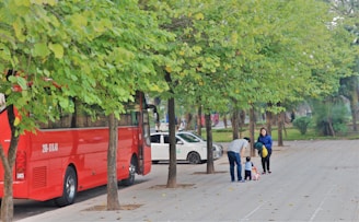 A red bus is parked along a tree-lined street with green leaves. Nearby, a man bends down to assist a small child on a scooter while a woman stands close by, smiling. A white car is parked further down the street.