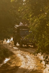 A rugged vehicle travels on a muddy and uneven dirt path surrounded by dense foliage. Several people are seated on top of the vehicle, enjoying the natural surroundings. The scene is bathed in a warm, golden light, suggesting a setting sun.