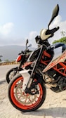 Two motorcycles are parked along a road with a scenic backdrop. The motorcycles have prominent orange and white coloring, with a focus on the tires and handlebars. The open road curves away, lined with a yellow and black barrier. In the background, there are hills and a clear sky.