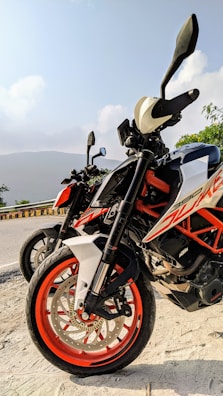 Two motorcycles are parked along a road with a scenic backdrop. The motorcycles have prominent orange and white coloring, with a focus on the tires and handlebars. The open road curves away, lined with a yellow and black barrier. In the background, there are hills and a clear sky.