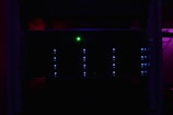 A dark server rack illuminated by faint blue LED lights, with a prominent green light in the center. The surrounding environment is dimly lit, creating a mysterious and technical atmosphere.
