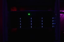 A dark server rack illuminated by faint blue LED lights, with a prominent green light in the center. The surrounding environment is dimly lit, creating a mysterious and technical atmosphere.