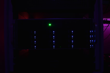 A dark server rack illuminated by faint blue LED lights, with a prominent green light in the center. The surrounding environment is dimly lit, creating a mysterious and technical atmosphere.