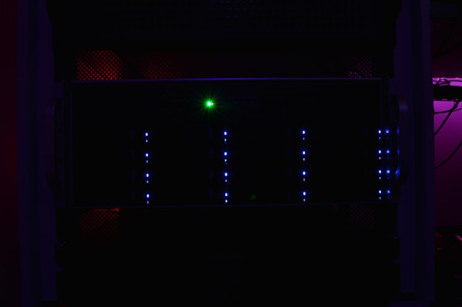 A dark server rack illuminated by faint blue LED lights, with a prominent green light in the center. The surrounding environment is dimly lit, creating a mysterious and technical atmosphere.