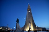 A large, modern church building with a distinctive, tall structure and illuminated entrance. The sky is dark blue, indicating evening or nighttime. A statue stands on the left side in front of the church, and several people are near the entrance.