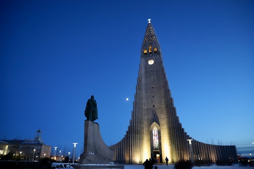 A large, modern church building with a distinctive, tall structure and illuminated entrance. The sky is dark blue, indicating evening or nighttime. A statue stands on the left side in front of the church, and several people are near the entrance.