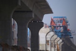 A construction site with large concrete pillars supporting an elevated structure. In the background, there is a blue and orange industrial crane with a number 3 visible on it. The setting appears urban, with buildings surrounding the construction area.