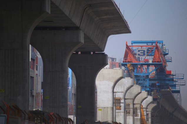 A construction site with large concrete pillars supporting an elevated structure. In the background, there is a blue and orange industrial crane with a number 3 visible on it. The setting appears urban, with buildings surrounding the construction area.