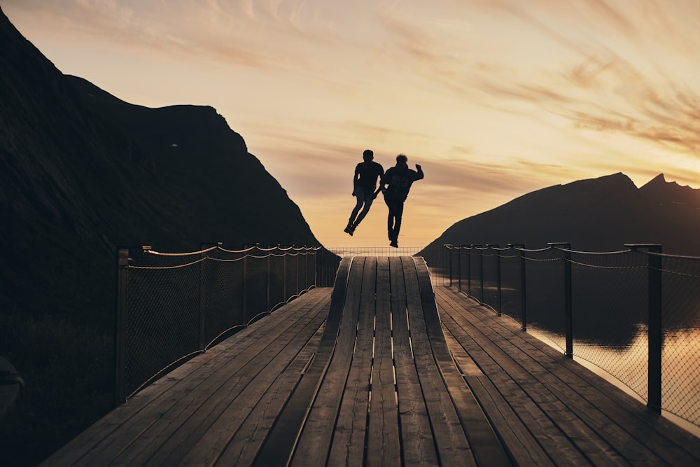silhouette view of couple jump on wooden hump beside body of water