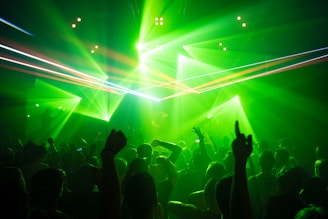 A large crowd of people is gathered in a dimly lit venue, enjoying a lively party or concert. Bright green laser lights and beams cut through the darkness, creating an energetic and exciting atmosphere. The silhouettes of people with raised hands suggest dancing or cheering, contributing to the vibrant scene.