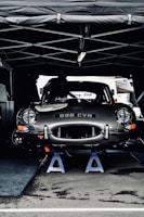 A vintage car with the license plate number 898 CVN is elevated on blue jack stands under a tent. The car appears to be in a service area, possibly during a motor event or race. Its sleek, shiny body and classic design are prominent.