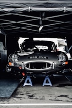 A vintage car with the license plate number 898 CVN is elevated on blue jack stands under a tent. The car appears to be in a service area, possibly during a motor event or race. Its sleek, shiny body and classic design are prominent.