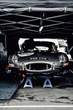 A vintage car with the license plate number 898 CVN is elevated on blue jack stands under a tent. The car appears to be in a service area, possibly during a motor event or race. Its sleek, shiny body and classic design are prominent.
