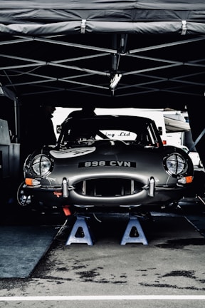 A vintage car with the license plate number 898 CVN is elevated on blue jack stands under a tent. The car appears to be in a service area, possibly during a motor event or race. Its sleek, shiny body and classic design are prominent.