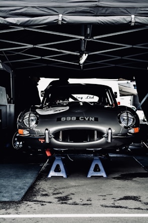 A vintage car with the license plate number 898 CVN is elevated on blue jack stands under a tent. The car appears to be in a service area, possibly during a motor event or race. Its sleek, shiny body and classic design are prominent.
