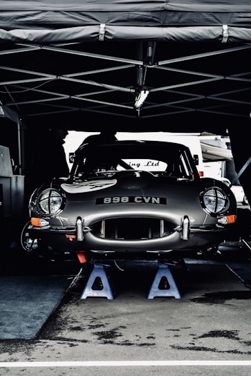 A vintage car with the license plate number 898 CVN is elevated on blue jack stands under a tent. The car appears to be in a service area, possibly during a motor event or race. Its sleek, shiny body and classic design are prominent.