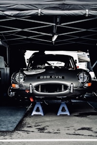 A vintage car with the license plate number 898 CVN is elevated on blue jack stands under a tent. The car appears to be in a service area, possibly during a motor event or race. Its sleek, shiny body and classic design are prominent.