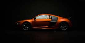 A sleek orange sports car is positioned sideways on a dark background, showcasing its aerodynamic design and stylish contours. The lighting highlights the glossy finish and the detailed features of the vehicle, including its alloy wheels and distinct side panel accents.