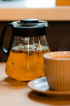 A glass teapot filled with herbal tea, featuring bits of herbs and lemon slices, sits on a wooden table next to a white ceramic cup with a matching saucer. The steam rising from the cup suggests that the tea is freshly brewed.