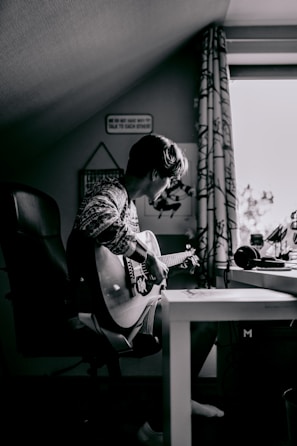 A person sits on a chair in a cozy room, playing an acoustic guitar. The setting is an attic or a small room with a sloped ceiling. The light comes from a nearby window, casting a soft glow. On the table beside the person, there are headphones and some other items. The scene conveys a sense of solitude and creativity.