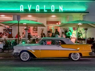 A classic yellow and white car is parked in front of an illuminated restaurant or cafe named 'AVALON' with a neon green sign. The establishment is bustling with people dining under large green umbrellas. The ambiance is vibrant and lively with customers sitting at tables and a band playing music inside.