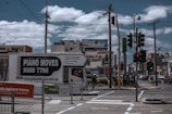 A busy urban street corner with a truck labeled 'Piano Moves' parked near pedestrian crossing signs. Several people stand at the crosswalk, waiting to cross. Overhead, power lines intersect the cloudy sky. In the background, a variety of shops and a billboard advertising an internet bundle are visible.