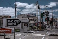 A busy urban street corner with a truck labeled 'Piano Moves' parked near pedestrian crossing signs. Several people stand at the crosswalk, waiting to cross. Overhead, power lines intersect the cloudy sky. In the background, a variety of shops and a billboard advertising an internet bundle are visible.
