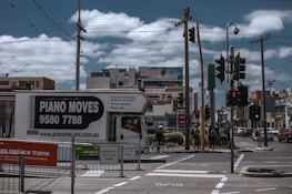 A busy urban street corner with a truck labeled 'Piano Moves' parked near pedestrian crossing signs. Several people stand at the crosswalk, waiting to cross. Overhead, power lines intersect the cloudy sky. In the background, a variety of shops and a billboard advertising an internet bundle are visible.