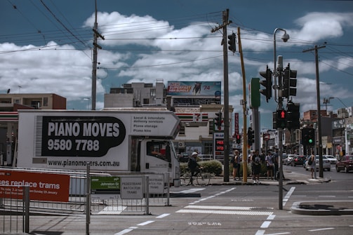A busy urban street corner with a truck labeled 'Piano Moves' parked near pedestrian crossing signs. Several people stand at the crosswalk, waiting to cross. Overhead, power lines intersect the cloudy sky. In the background, a variety of shops and a billboard advertising an internet bundle are visible.