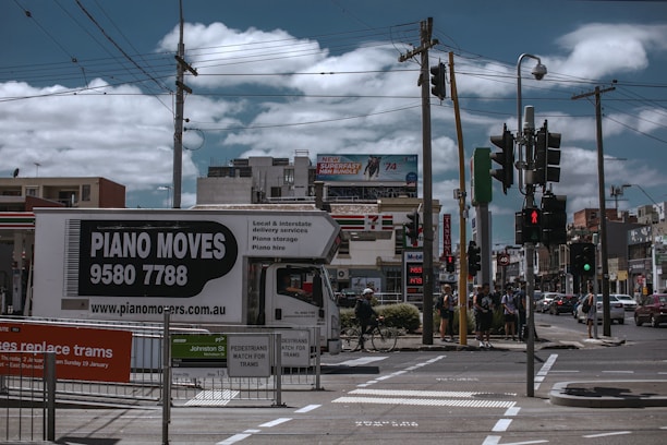 A busy urban street corner with a truck labeled 'Piano Moves' parked near pedestrian crossing signs. Several people stand at the crosswalk, waiting to cross. Overhead, power lines intersect the cloudy sky. In the background, a variety of shops and a billboard advertising an internet bundle are visible.