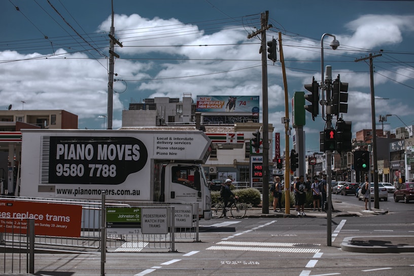 A busy urban street corner with a truck labeled 'Piano Moves' parked near pedestrian crossing signs. Several people stand at the crosswalk, waiting to cross. Overhead, power lines intersect the cloudy sky. In the background, a variety of shops and a billboard advertising an internet bundle are visible.